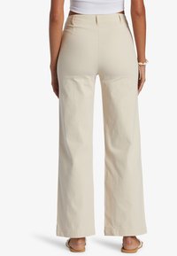 Frau in cremefarbenen weit geschnittenen Hosen, weißem Cropped-Top und weißen Sandalen, die mit dem Rücken zu einer schlichten weißen Wand steht.