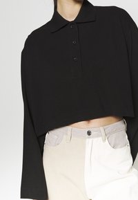 Person mit einem langärmligen, schwarzen, kurzen Polohemd und hochgeschnittenen, beige-weißen, farblich abgesetzten Jeans, sichtbarem Bauch.