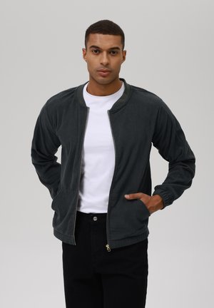 Dark green corduroy bomber jacket with a zip front, ribbed collar, and elastic wristbands; paired with a white crew-neck shirt and black pants.