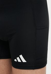Shorts de sport noirs avec poche latérale et logo Adidas blanc portés sur la partie supérieure de la cuisse.