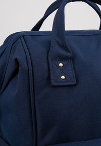 Námořnický modrý látkový batoh s dvojitými horními uchy, texturovaným materiálem, zesíleným šitím a zlatými kovovými detaily.