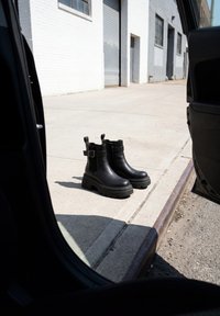 Black leather boots with thick soles placed on sidewalk next to open car door and curb in an urban area with white buildings.