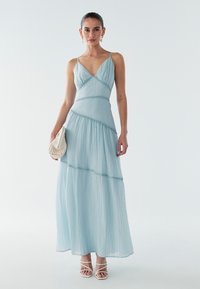 Světlomodré maxi šaty s výstřihem do V, tenkými ramínky, vrstvenou látkou a texturovanými detaily. Kombinované s malou pletenou kabelkou a sandály na tenkých páscích.