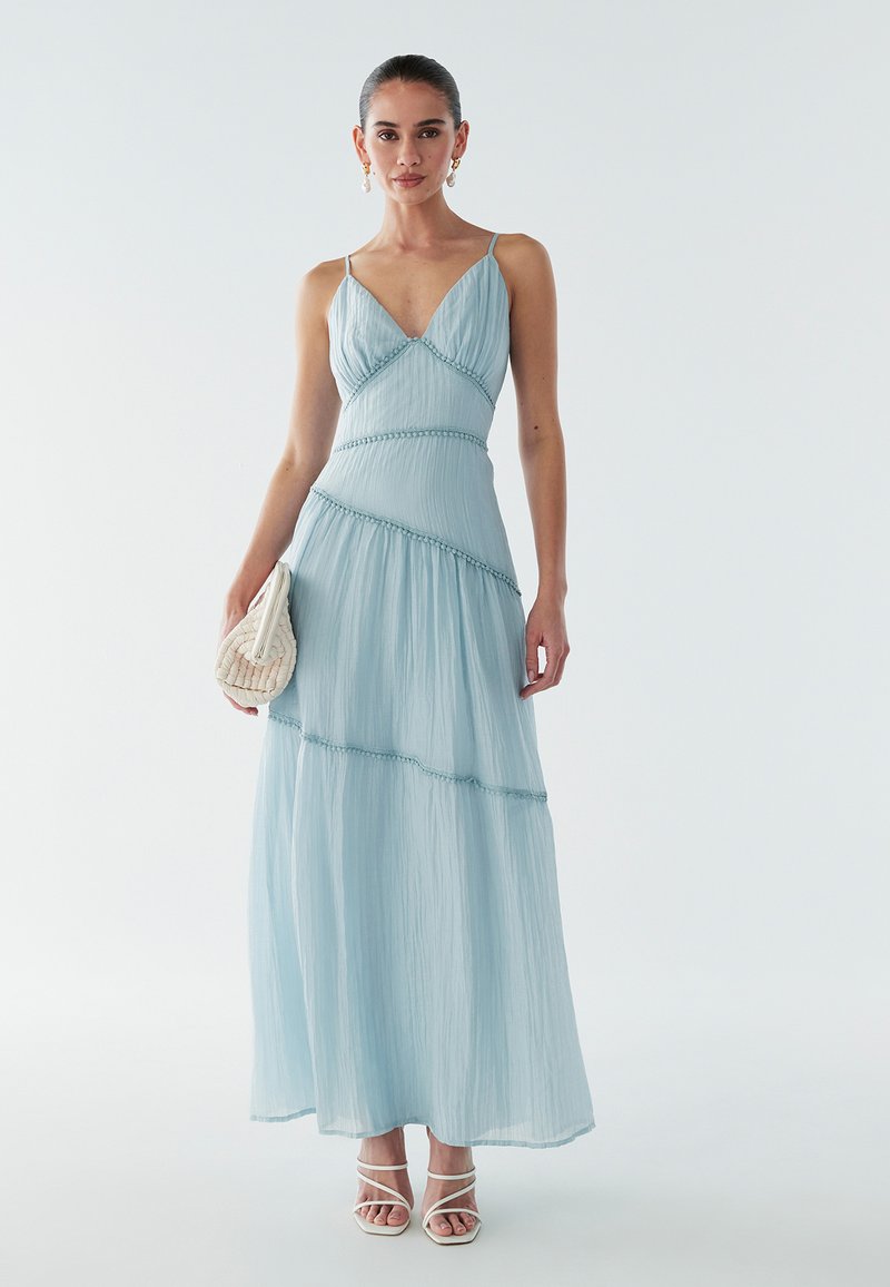 Světlomodré maxi šaty s výstřihem do V, tenkými ramínky, vrstvenou látkou a texturovanými detaily. Kombinované s malou pletenou kabelkou a sandály na tenkých páscích.