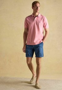 Rozā īsām piedurknēm polo krekls ar mazu zilu logo, kopā ar zilām šortiem, kuros ir bāka raksts, un gaiši brūnām slip-on kurpēm.