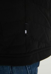 Schwarzer Steppjackensaum mit kleinem Stoffetikett oberhalb von unten sichtbar blauer Jeans.