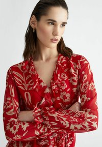 Red floral blouse featuring a ruffled neckline and cuffs, with a sheer texture and gold floral patterns. Long sleeves with elastic cuffs.