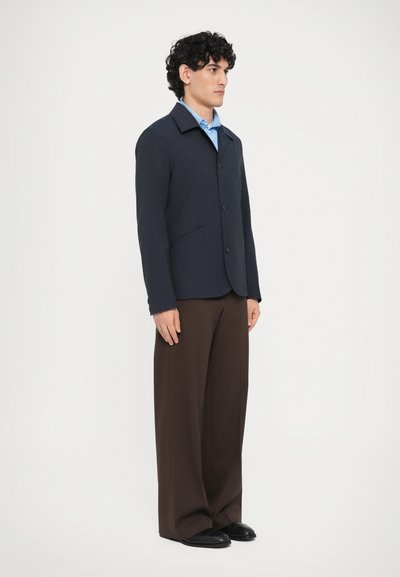 Tengerészkék gyapjú zakó felvágott gallérral, elülső gombos záródással és oldalzsebekkel, laza barna nadrággal és fekete cipővel párosítva.