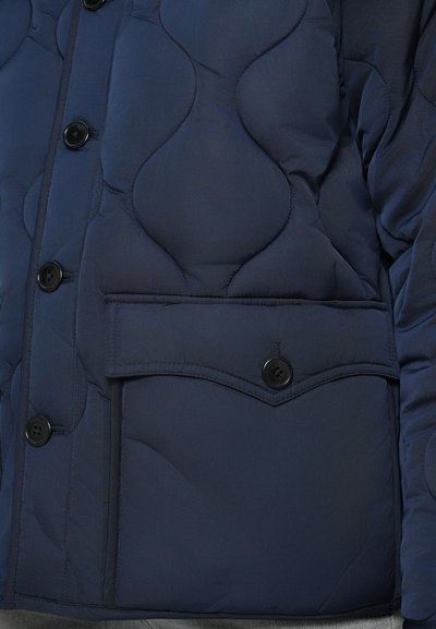 Marineblå quilted jakke med rundede mønstre, sorte knapper og en klaplomme med buet kant på venstre side. Struktureret stof.