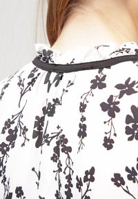 Svart blommönster på en vit tygblus med volangkrage och svart kant, med flödande, lätt textur.