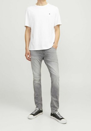 Hombre con camiseta blanca lisa de manga corta, vaqueros ajustados de color gris claro y zapatillas bajas en blanco y negro, de pie frente a un fondo liso.