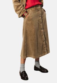 Jupe midi en velours côtelé marron avec un devant boutonné, des poches latérales et une texture en côtes verticales. Associée à des chaussettes grises et des mocassins foncés.