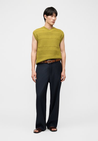 Jeune homme portant un haut vert en tricot sans manches, un pantalon large à fines rayures bleu marine, une ceinture marron et des sandales foncées, debout devant un fond uni.