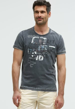 Mann trägt ein ausgewaschenes dunkelgraues Grafik-T-Shirt und hellbeige Cargo-Shorts, steht mit entspannten Händen an den Seiten.