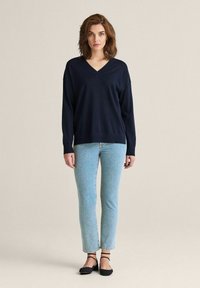 Marinblå v-ringad tröja i mjukt tyg, avslappnad passform, tillsammans med ljusblå raka jeans och svarta lågskor, står på en neutral bakgrund.