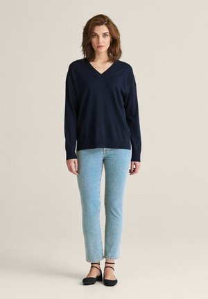 Marineblå v-hals sweater i blødt materiale, afslappet pasform, parret med lyseblå lige bukser og sorte flade sko, stående på en neutral baggrund.