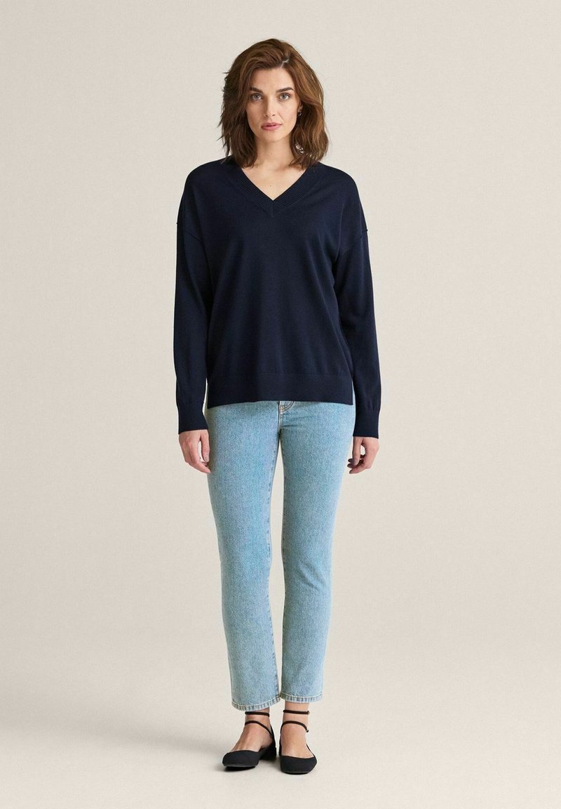 Marinblå v-ringad tröja i mjukt tyg, avslappnad passform, tillsammans med ljusblå raka jeans och svarta lågskor, står på en neutral bakgrund.