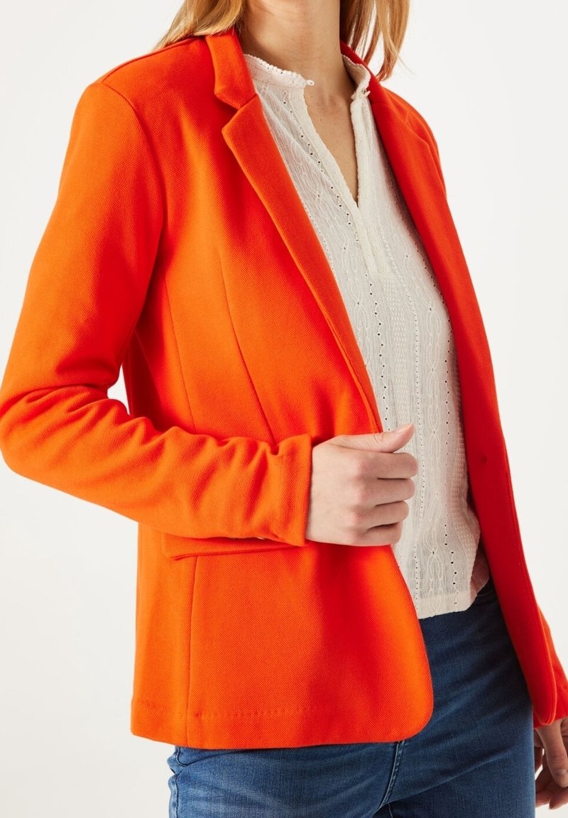 Femme portant un blazer orange vif sur un chemisier blanc texturé et un jean bleu, ajustant le devant du blazer d'une main.