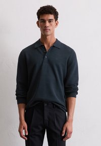 Dunkelgrünes Langarm-Polohemd mit strukturiertem Muster, Knopfleiste und gerippten Bündchen, kombiniert mit schwarzen Hosen.
