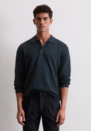 Dark green long-sleeve polo shirt with a textured pattern, buttoned placket, and ribbed cuffs, paired with black trousers.