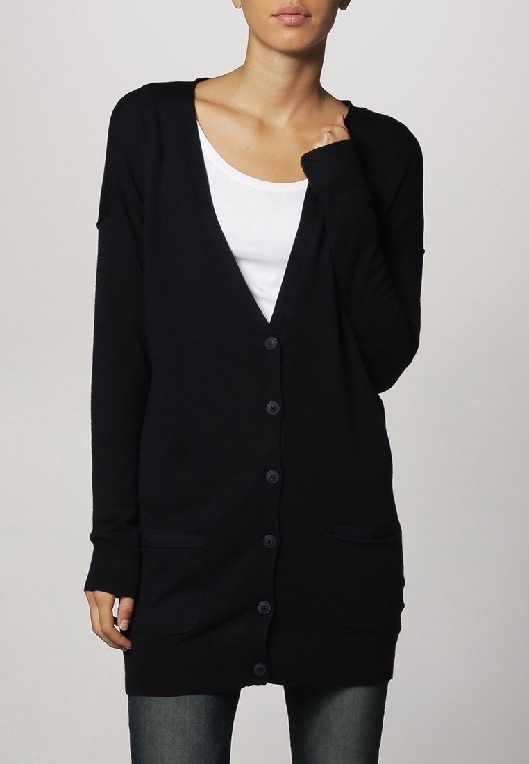 Femme portant un long cardigan noir boutonné avec des poches avant, sur un haut blanc et un jean bleu, debout devant un fond uni.