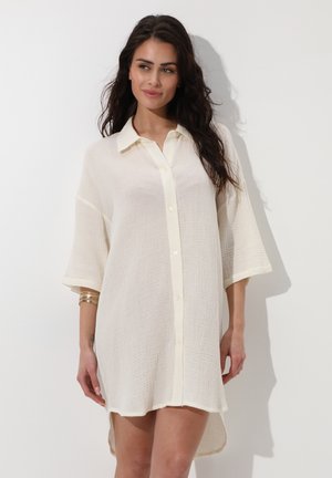 Donna con lunghi capelli scuri che indossa un vestito camicia sciolto e testurizzato di colore bianco sporco con bottoni e maniche a tre quarti, in piedi davanti a uno sfondo bianco.