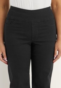 Nainen, jolla on ylipitkät mustat housut ja valkoinen toppi, kädet sivuilla, kuvattuna vyötäröstä reisiin.