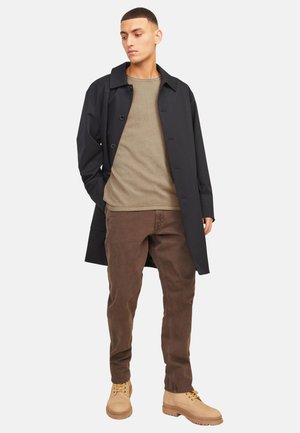 Junger Mann, der einen schwarzen Mantel über einem beigen Pullover, braune Hose und hellbraune Stiefel trägt, steht mit den Händen in den Taschen und blickt zur Seite.