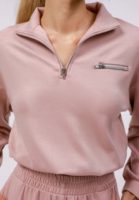 Pullover rose clair à demi-zip en tissu doux avec une fermeture éclair argentée. Il dispose d'une poche poitrine avec une fermeture éclair assortie et d'une taille froncée.