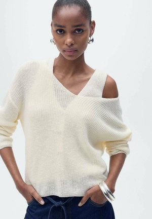 Light cream sweater with a V-neck and one shoulder exposed, featuring a loose knit texture. Paired with dark denim jeans and silver bracelets.