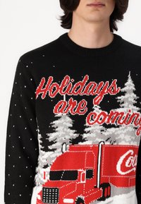 Čierny pletený sveter s vianočným motívom nákladného auta v červenej farbe, bielych stromoch a textom "Sviatky prichádzajú" v červenej a bielej farbe.