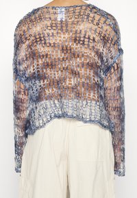 Personne portant un pull en maille ample à motif quadrillé bleu et blanc sur un pantalon beige, vue de dos sur un fond uni.