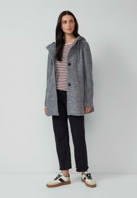 Manteau en laine gris avec capuche, comportant des boutons et des poches, porté par-dessus un haut rayé, assorti à un pantalon foncé et des chaussures sportives.