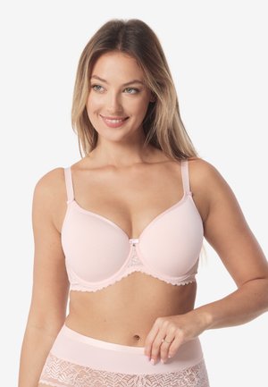 Kobieta o długich blond włosach, ubrana w jasnoróżowy, pikowany biustonosz i dopasowane koronkowe majtki, uśmiechająca się i pozująca na białym tle.