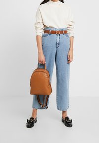 Personne portant un pull crème et un jean taille haute bleu clair, tenant un sac à dos en cuir marron, des mocassins noirs avec des boucles dorées.