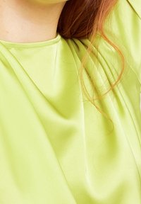 Světle zelený saténový top s jemným drapováním, kulatým výstřihem a jemnou texturou. Na látce je vidět pramínek vlasů.