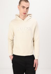 Sudadera beige con un bolsillo frontal y cordón. Presenta un logo de "GUESS" bordado en plata y un acento de "EST. 1981". Material suave y liso.