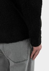 Sort tekstureret sweater med en knappet mansjet, båret over lysegr�å bukser med en baglomme. Et nærbillede af tøjets detaljer.