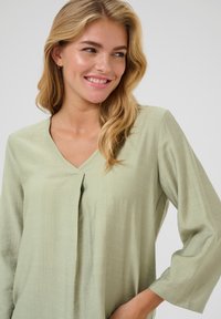 Vaaleanvihreä pitkähiha blouse, jossa on V-pääntie ja pleat-dekoratiivinen yksityiskohta rinnassa. Valmistettu tekstuurisesta, pehmeästä kankaasta.