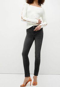 Nainen, joka on pukeutunut valkoiseen pitkähiharaiseen paitaan, jossa on hihaleikkaukset, mustiin muihin farkkuihin ja oransseihin korkokenkiin, seisoo tasaisella taustalla.