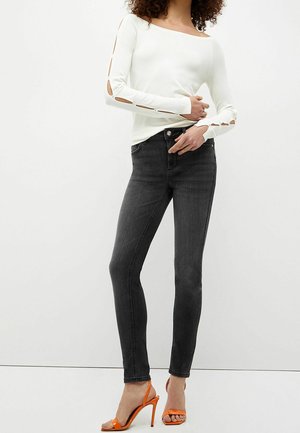 Mujer vestida con una blusa blanca de manga larga con cortes en los brazos, jeans ajustados negros y sandalias de tacón alto naranjas, de pie contra un fondo sencillo.