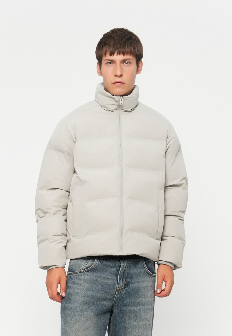 Graue Pufferjacke mit hohem Kragen, mit einem Reißverschluss vorne und gestepptem Design. Kombiniert mit blauen Jeans.
