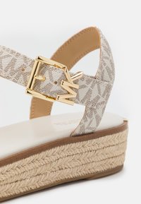 Gros plan d'une sandale compensée beige avec une semelle en corde tressée et une bride à motifs ornée d'une boucle dorée avec des initiales décoratives.