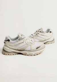 Baskets blanches et beiges avec des superpositions en mesh et synthétique, fermeture à lacets, col rembourré et semelle en caoutchouc texturée avec motifs antidérapants.