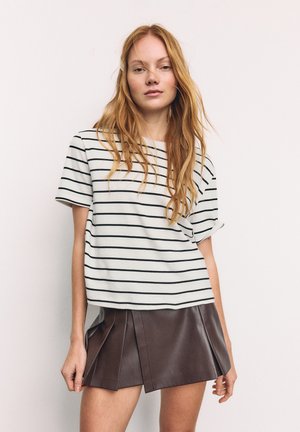 Young woman with long red hair wearing a white and black striped t-shirt and a brown pleated leather skirt, standing against a plain background.