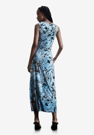 Mujer con el cabello trenzado que lleva un vestido maxi sin mangas de color azul claro con patrones florales negros, de pie con una mano en la cadera y tacones negros.