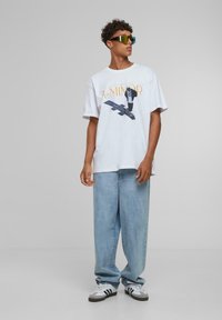 Hvit grafisk t-skjorte med teksten "A-MINOR", kombinert med lyseblå baggy jeans. Modellen har på seg grønn-tonede solbriller og hvite joggesko.