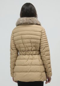 Donna con lunghi capelli scuri che indossa un giubbotto imbottito beige con cintura elastica e colletto di pelliccia, vista da dietro.