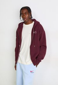 Bordo jaciņa ar rāvējslēdzēju un priekšējām kabatām, kombinēta ar gaiši bežīgu T-kreklu un gaiši ziliem sporta biksēm ar sarkano Nike logotipu un tekstu.