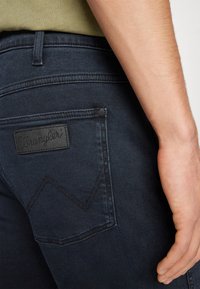 Zadnji žep temno modrih Wrangler jeansov, ki jih nosi oseba v bež majici, prikazuje usnjeno oznako blagovne znamke in roko, ki počiva ob boku.